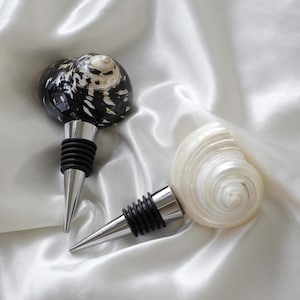 Op de afbeelding: Twee decoratieve flessenstoppers gemaakt van schelpen. Eén is zwart-wit met een zilveren bovenkant. De andere is wit met een zilveren bovenkant.