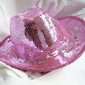 May include: A pink cowboy hat covered in small, reflective squares, creating a mosaic effect. The hat has a classic cowboy hat shape with a wide brim and a slightly curved crown. The brim and crown are outlined with a pink beaded trim.