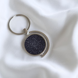 May include: A silver-colored keychain with a circular pendant featuring a dark blue background and a white constellation design. The keychain is made of metal and has a ring for attaching keys.