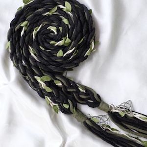 May include: Black, green, and white braided yarn with green leaves woven throughout. The yarn is tied together with silver Celtic knot charms.