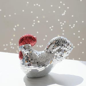 May include: A decorative rooster figurine covered in mirrored tiles. The rooster has a red comb and wattle, with the rest of its body covered in small, reflective squares. The figurine is positioned on a white surface.