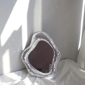 Puede incluir: Un espejo plateado en forma de corazón con un diseño de mosaico. El espejo está hecho de pequeñas baldosas reflectantes que crean un efecto brillante.