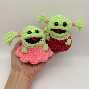 May include: Two crocheted plush toys with green bodies, pink dresses, and black eyes. The toys have pink yarn pigtails and red yarn mouths.
