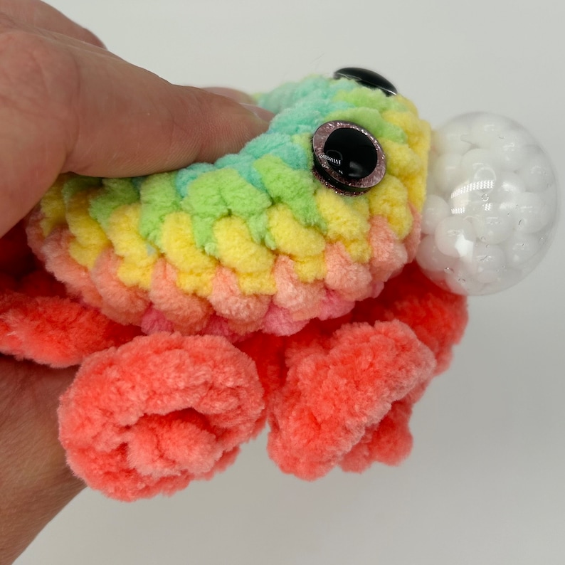 May include: A rainbow colored crocheted octopus with a clear plastic sphere filled with white beads. The octopus has black eyes with pink glitter accents.