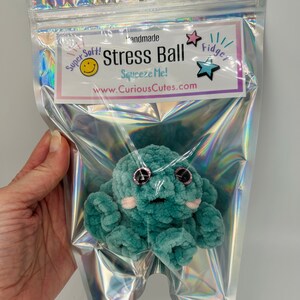 May include: A teal blue crocheted octopus stress ball with pink cheeks and black eyes. The octopus is in a clear plastic bag with a label that reads "Handmade Stress Ball Squeeze Me! www.CuriousCutes.com" and a star with the word "Fidget" on it.