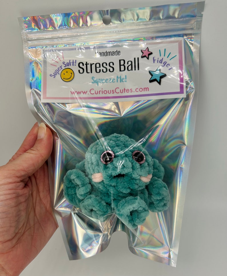 May include: A teal blue crocheted octopus stress ball with pink cheeks and black eyes. The octopus is in a clear plastic bag with a label that reads "Handmade Stress Ball Squeeze Me! www.CuriousCutes.com" and a star with the word "Fidget" on it.