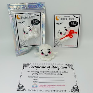 Könnte beinhalten: Ein handgefertigtes weißes Häkelgespenst mit schwarzen Augen und rosa Rouge, mit dem Text "Hi, Boo!" in einer Sprechblase. Das Gespenst ist in einer holografischen Tüte und auf einer Karte verpackt. Ein Adoptionszertifikat ist ebenfalls enthalten.