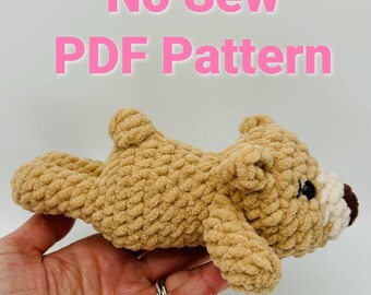 May include: A crocheted brown teddy bear with white and brown accents. The bear is lying on its side with its head turned to the right. The text "No Sew PDF Pattern" is written in pink letters above the bear.