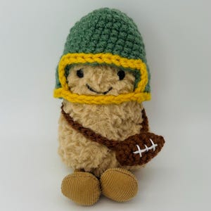 May include: A plush toy shaped like a tan, fuzzy creature wearing a green and yellow crocheted helmet. The figure has black bead eyes, a stitched smile, and a brown crocheted football-shaped bag with white laces hanging across its body.