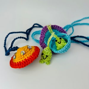 May include: A crocheted necklace with a rainbow-colored UFO charm and two green alien charms. The UFO charm has rhinestones on it.