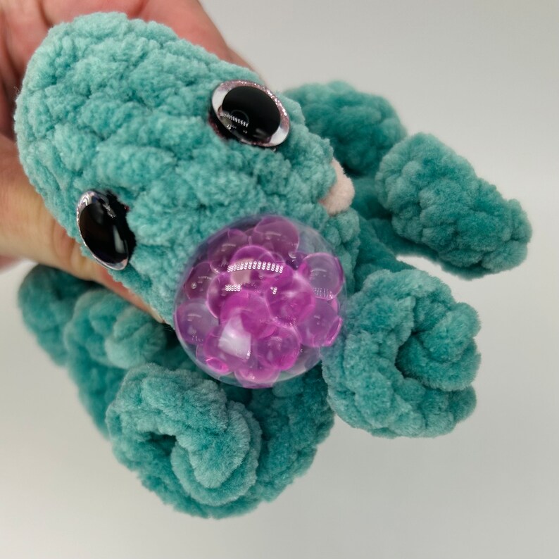 May include: A teal blue crocheted octopus plush toy with large black eyes and a clear plastic bubble filled with pink beads.