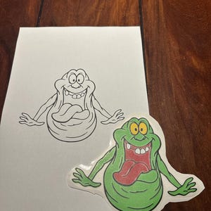May include: A black and white line drawing of a green ghost with a large tongue sticking out. The ghost is on a white sheet of paper. Below the drawing is a green, red, and yellow sticker of the same ghost.