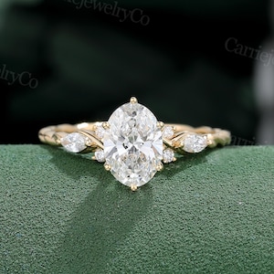 May include: A gold engagement ring with a large oval-cut diamond center stone. The ring features smaller diamonds and a detailed band. The ring is set against a green background.
