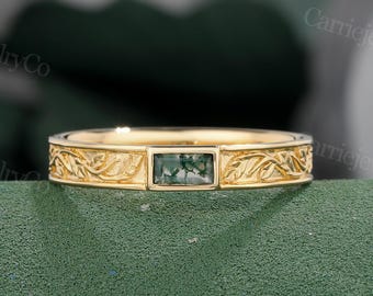Puede incluir: Un anillo de oro con una piedra verde rectangular en el centro. La banda presenta un intrincado diseño floral. El anillo se muestra sobre una superficie verde, mostrando los detalles de la joya.