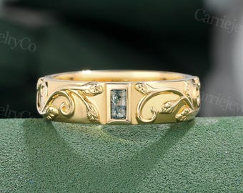 Puede incluir: Un anillo dorado con un diseño floral grabado y una gema rectangular de color verde claro. La banda del anillo presenta patrones detallados de hojas y vides. La gema está engastada en el centro.