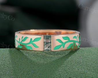Puede incluir: Un anillo dorado con una gema verde rectangular en el centro. La banda presenta un diseño de hojas verdes vibrantes. El anillo se muestra sobre un fondo verde, resaltando el diseño de la joya.