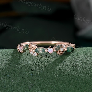 May include: A delicate rose gold band with a floral design featuring marquise-shaped green gemstones and round white diamonds. Two oval-shaped opals are set in the center of the band.