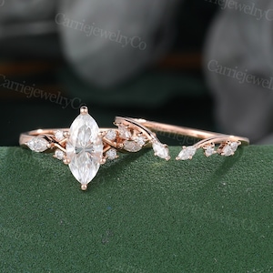 May include: A rose gold engagement ring set. The engagement ring features a large marquise-cut diamond centre stone, surrounded by smaller diamonds and leaf-shaped accents. The matching band has a curved design with additional diamonds.