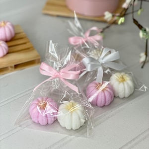 May include: Four small pumpkin-shaped candles, two white and two pink, are packaged in clear cellophane bags with pink and white ribbons. The candles have a gold glitter accent.