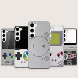 Könnte beinhalten: Eine Sammlung von fünf Handyhüllen mit Retro-Videospielkonsolen-Designs. Die Hüllen zeigen Designs, die von klassischen Konsolen inspiriert sind, darunter eine PlayStation, ein Game Boy und andere Controller. Die Hüllen sind in verschiedenen Farben erhältlich, darunter Grau, Schwarz und Weiß.