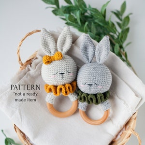 May include: Two crocheted bunny rattle toys with wooden rings. One bunny is gray with green trim and the other is gray with yellow trim. The bunnies are in a wicker basket with a white cloth.