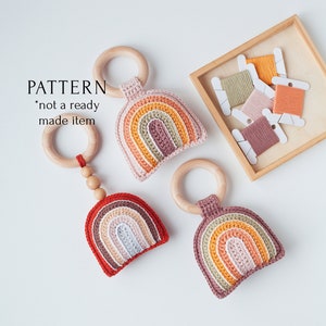 May include: Three crocheted rainbow teethers with wooden rings. The teethers are in shades of pink, orange, and red. The wooden rings are attached to the teethers with wooden beads. The teethers are on a white surface.  PATTERN *not a ready made item*