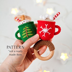 May include: A crocheted Christmas-themed toy with two mugs, one green and one red, attached to a wooden ring. The green mug has a Christmas tree design and the red mug has a snowflake design. The toy is perfect for a baby's first Christmas.