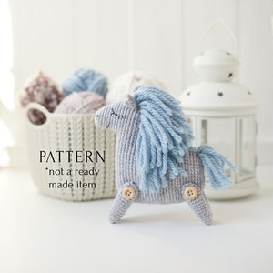 Puede incluir: Un caballo gris de crochet con una crin y una cola azules. El caballo tiene botones de madera para los ojos y está sentado sobre una superficie blanca. El texto "PATTERN *not a ready made item" es visible en la imagen.
