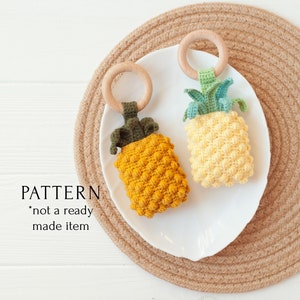 Könnte beinhalten: Zwei gehäkelte Baby-Spielzeuge in Ananasform mit Holzringen, eines in Gelb und das andere in Senf. Die Spielzeuge liegen auf einem weißen Teller auf einer gewebten Matte. Der Text "PATTERN *not a ready made item" ist sichtbar.