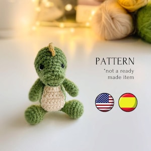 May include: Green crocheted dinosaur amigurumi toy with a white belly. The toy is sitting on a white surface. The text "PATTERN *not a ready made item*" is visible in the image. The American and Spanish flags are also visible.