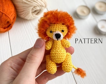 May include: A crocheted lion amigurumi toy with a yellow body, orange mane, and black eyes. The toy is being held in a hand. The word "PATTERN" is written in white text on a white background.