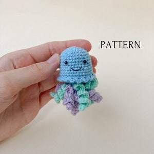 May include: A crocheted jellyfish toy with a light blue body, purple and green tentacles, and a smiling face. The toy is being held in a hand.