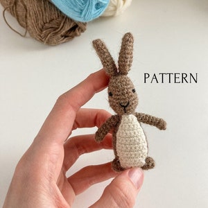May include: A small crocheted brown bunny with white accents. The bunny is being held in a hand. The word "PATTERN" is written in white text on a white background.