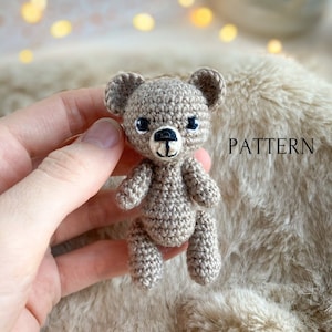 May include: A small crocheted brown teddy bear with black eyes and a black nose. The bear is sitting on a fluffy beige surface. PATTERN is written in white text on the surface.