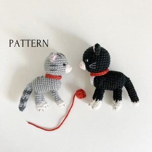Peut inclure: Deux jouets en crochet en forme de chat, l'un gris et blanc et l'autre noir et blanc, avec des colliers en laine rouge. Le chat gris et blanc tient une pelote de laine rouge.