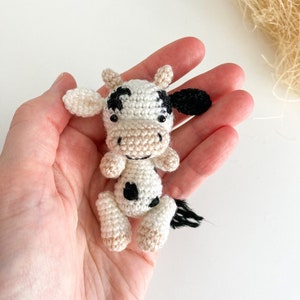 May include: A small crocheted cow toy with black and white spots. The cow is sitting in a hand, with its head tilted to the side.