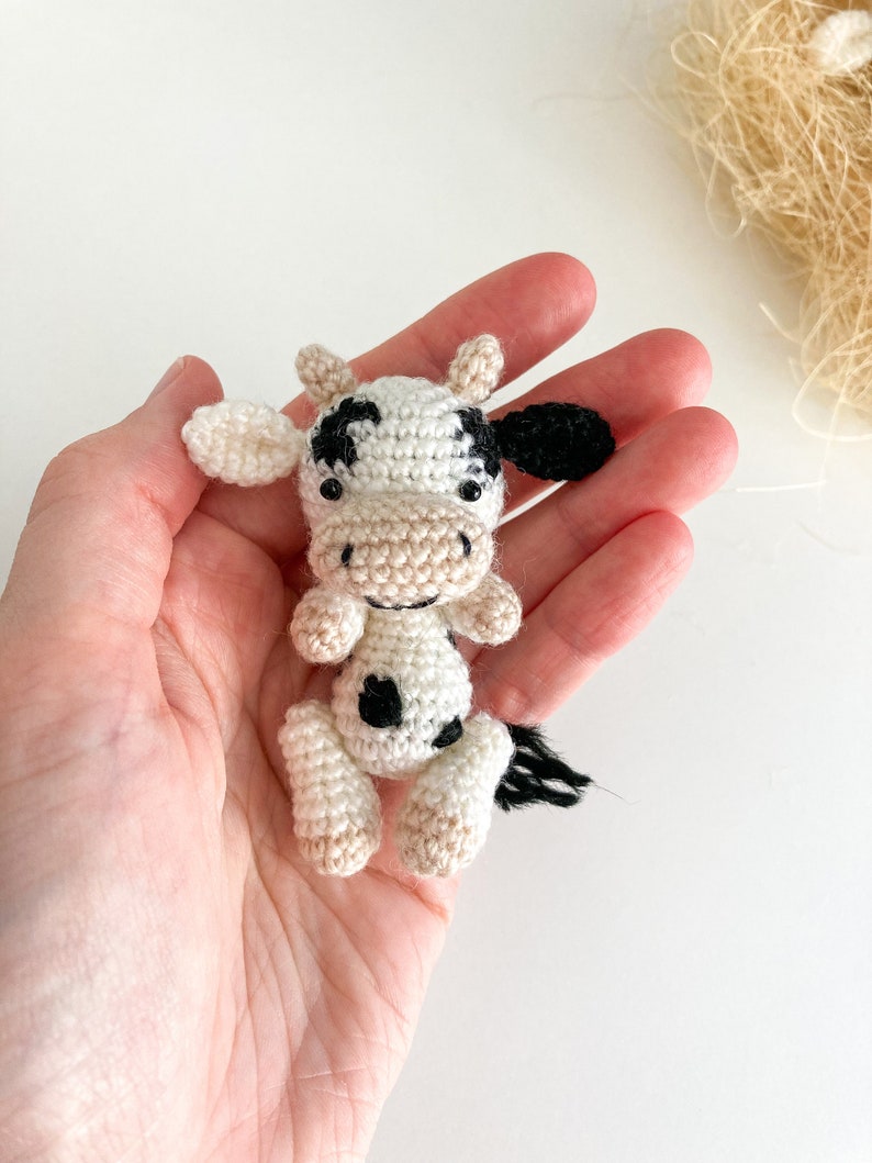 May include: A small crocheted cow toy with black and white spots. The cow is sitting in a hand, with its head tilted to the side.