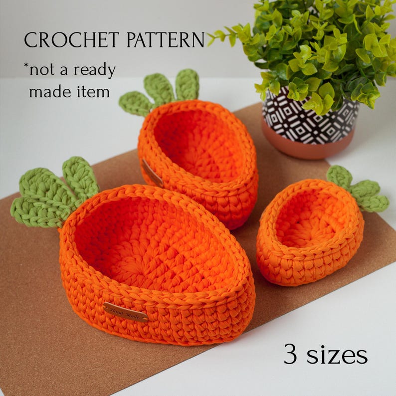 May include: Three crocheted carrot-shaped baskets in orange with green tops. The baskets are of different sizes. The text "CROCHET PATTERN" and "*not a ready made item" is visible in the image. The text "3 sizes" is also visible in the image.