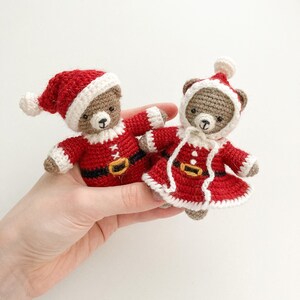K&ouml;nnte beinhalten: Zwei geh&auml;kelte Teddyb&auml;ren, die rote und wei&szlig;e Weihnachtsmannkost&uuml;me tragen. Die B&auml;ren halten sich an den H&auml;nden und sitzen auf einer wei&szlig;en Oberfl&auml;che.