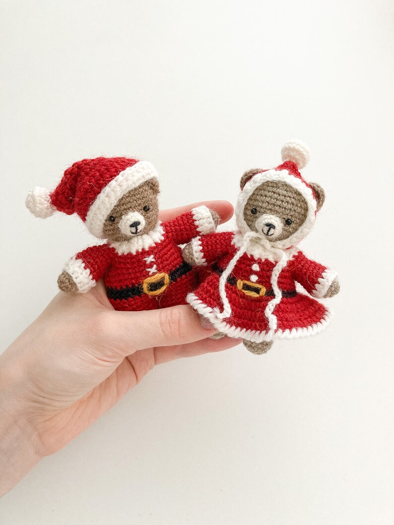 K&ouml;nnte beinhalten: Zwei geh&auml;kelte Teddyb&auml;ren, die rote und wei&szlig;e Weihnachtsmannkost&uuml;me tragen. Die B&auml;ren halten sich an den H&auml;nden und sitzen auf einer wei&szlig;en Oberfl&auml;che.