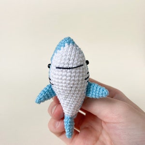 May include: A small crocheted shark toy with a white body and light blue fins and tail. The shark has black eyes and a black mouth.