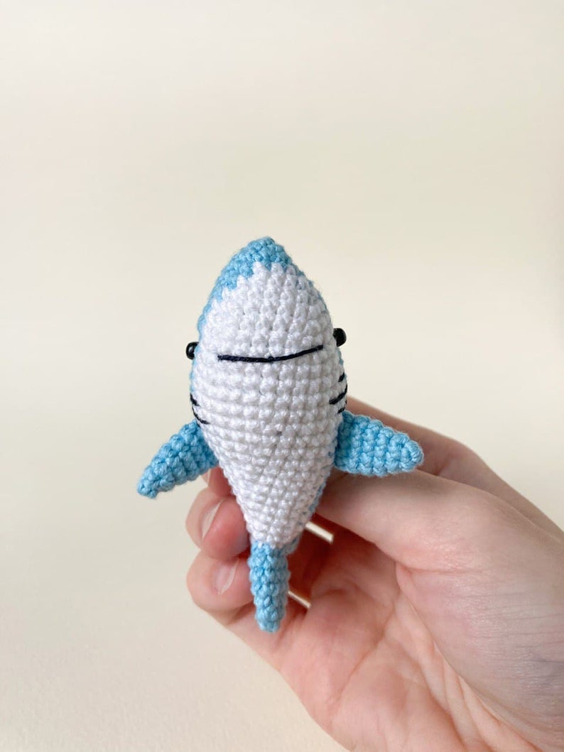 May include: A small crocheted shark toy with a white body and light blue fins and tail. The shark has black eyes and a black mouth.