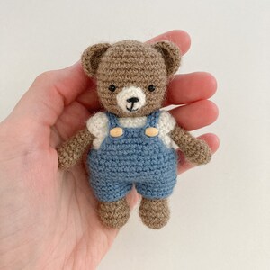 May include: A small brown crocheted teddy bear wearing blue overalls with white accents and wooden buttons. The bear is being held in a hand.