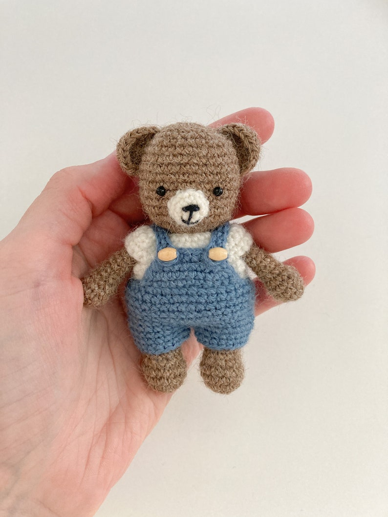 May include: A small brown crocheted teddy bear wearing blue overalls with white accents and wooden buttons. The bear is being held in a hand.