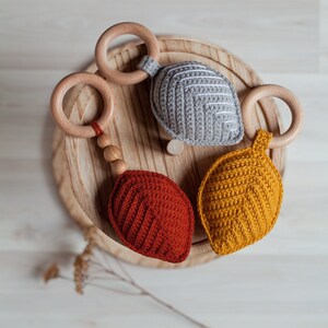 May include: Three crocheted leaf-shaped teethers with wooden rings. The teethers are in shades of gray, yellow, and red.