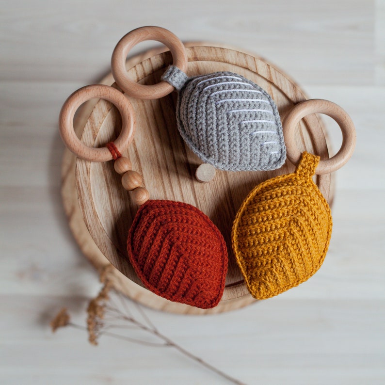May include: Three crocheted leaf-shaped teethers with wooden rings. The teethers are in shades of gray, yellow, and red.