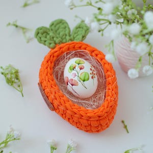 May include: A crocheted carrot-shaped Easter basket with a white egg decorated with floral designs inside. The basket is orange and green and is sitting on a white surface with white flowers.