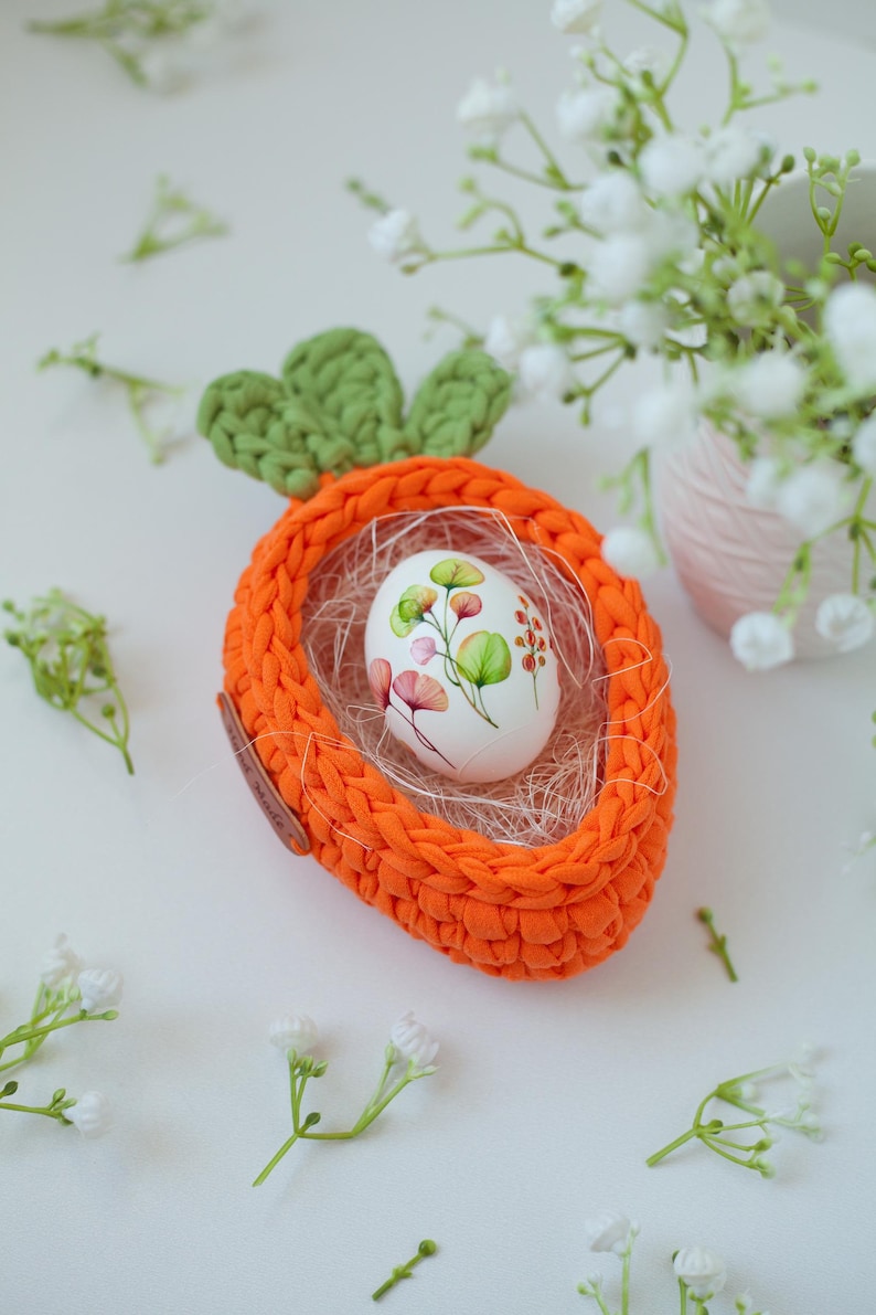 May include: A crocheted carrot-shaped Easter basket with a white egg decorated with floral designs inside. The basket is orange and green and is sitting on a white surface with white flowers.