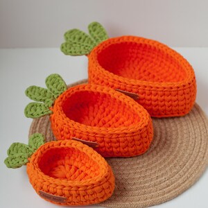 May include: Three crocheted carrot-shaped baskets in orange and green, with the smallest basket labeled "Hand Made", sitting on a woven brown mat.