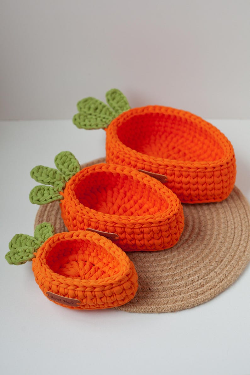 May include: Three crocheted carrot-shaped baskets in orange and green, with the smallest basket labeled "Hand Made", sitting on a woven brown mat.